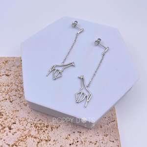 A pair of sterling silver dangling giraffe stud earrings displayed on a white stand against a textured background.